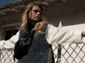 Une jeune femme porte un pull sans manche en maille bleue sur une chemise blanche, un sac banane noir et un bracelet doré. Elle s'appuie sur une clôture rouillée.