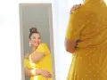 Une femme se regarde dans un miroir, souriante, portant une robe jaune à pois. Elle semble heureuse, malgré la robe trop courte.