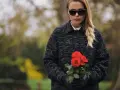 Une jeune femme, portant une tenue d'enterrement hiver, tient un bouquet de roses rouges. Ses lunettes de soleil cachent ses yeux, mais son expression est empreinte de tristesse.