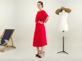 Une femme pose dans une robe rouge éclatante. À ses côtés, une chaise longue, un mannequin de couture et des plantes. Une robe casual, c'est quoi ?