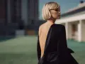 Une femme au style glamour, dos nu dans une robe noire satinée, pose devant un bâtiment moderne.