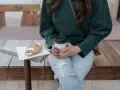 Une femme en pull vert, jean déchiré, savoure un café et un croissant. Parfait pour un look décontracté.