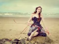 Jeune femme au style pin-up année 50, assise sur un rocher sur la plage, robe à pois, ruban rouge flottant au vent.