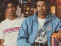 Deux jeunes hommes posent, arborant un style vestimentaire année 80 : t-shirt à rayures roses et veste bleue.