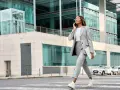 Femme en tenue d'entretien d'embauche d'été, tailleur gris, traversant la rue avec un ordinateur portable, parlant au téléphone.