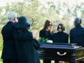 Un couple en tenue d'enterrement, le bras autour de l'épaule, se tient près d'un cercueil. D'autres personnes en deuil sont derrière.