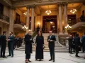 Trois personnes en tenue d'opéra discutent dans un grand hall orné de colonnes et d'escaliers majestueux.