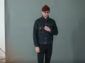 Homme portant une veste en jean sombre et un pantalon noir, bonnet bordeaux et lunettes rondes.