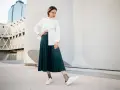 Une femme pose avec confiance, portant une chemise blanche et une élégante jupe plissée vert foncé.