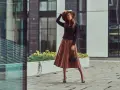 Une femme élégante porte une jupe longue plissée et des bottines à talons aiguilles, un chapeau à la main.