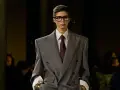 Mannequin au style collegienne, portant un costume gris oversize, chemise à rayures et cravate bordeaux. Lunettes imposantes.