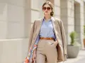 Une femme porte une chemise bleu avec un pantalon beige, un blazer camel et des lunettes de soleil.