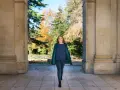 Une femme souriante marche sous une arche, portant un manteau vert et un haut bleu marine. L'automne colore les arbres en arrière-plan.