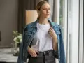 Une femme porte une veste en jean bleue sur un t-shirt blanc et un jean gris. Elle a des colliers superposés et des bagues.