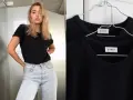 Une jeune femme porte une tenue avec t-shirt noir et jean clair. Deux t-shirts noirs identiques sont accrochés sur des cintres.
