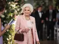 Élégante femme de 75 ans, radieuse dans sa tenue de mariage rose poudré, collier de perles et broche scintillante.