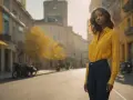 Une femme porte une chemise jaune oversize rentrée dans un jean. Elle se tient dans une rue bordée de bâtiments jaunes et d'arbres aux feuilles d'automne.