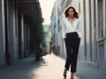 Une femme élégante marche dans une ruelle, portant une chemise blanche et un pantalon noir.