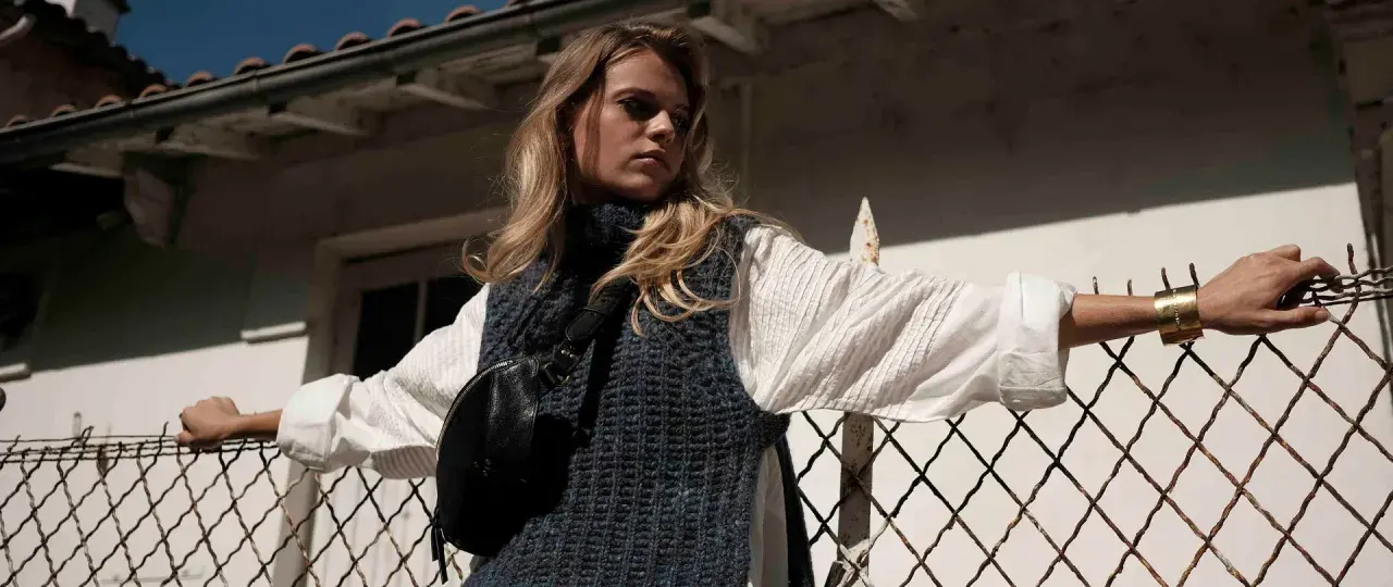 Une jeune femme porte un pull sans manche en maille bleue sur une chemise blanche, un sac banane noir et un bracelet doré. Elle s'appuie sur une clôture rouillée.