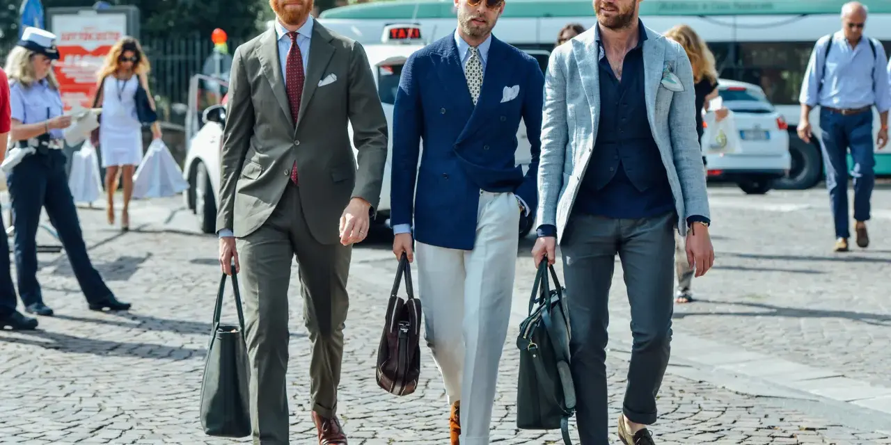 Trois hommes élégants déambulent. L'un porte une tenue avec blazer bleu ciel, un autre un costume vert olive, et le troisième un blazer gris clair.