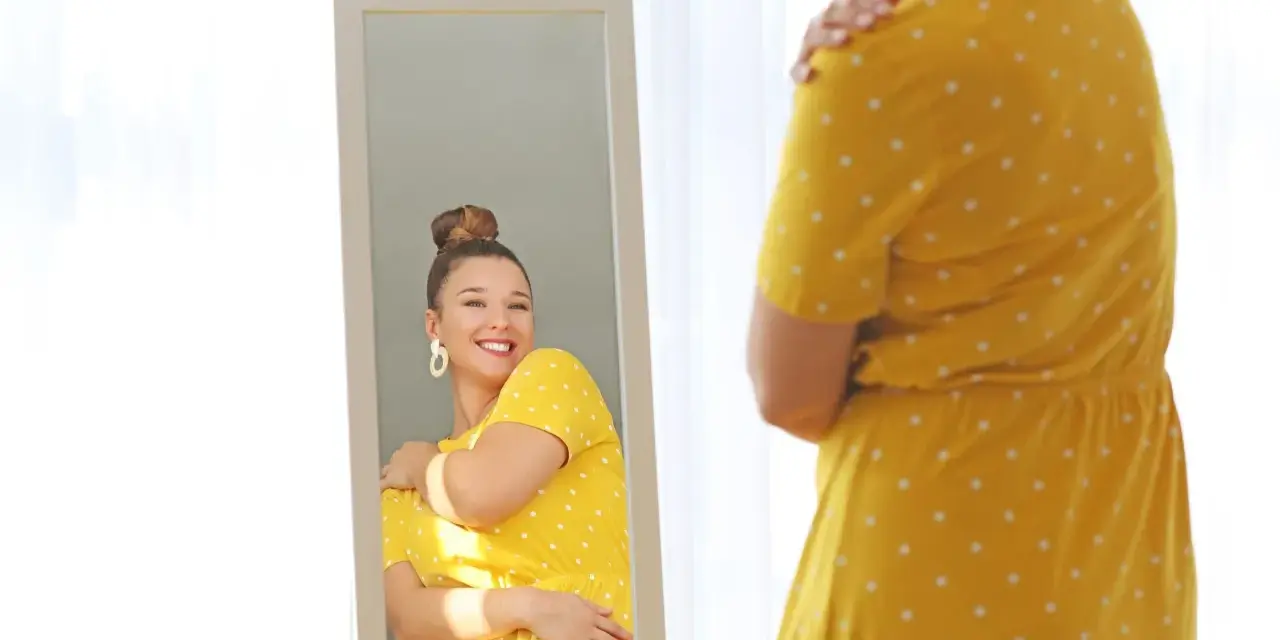 Une femme se regarde dans un miroir, souriante, portant une robe jaune à pois. Elle semble heureuse, malgré la robe trop courte.