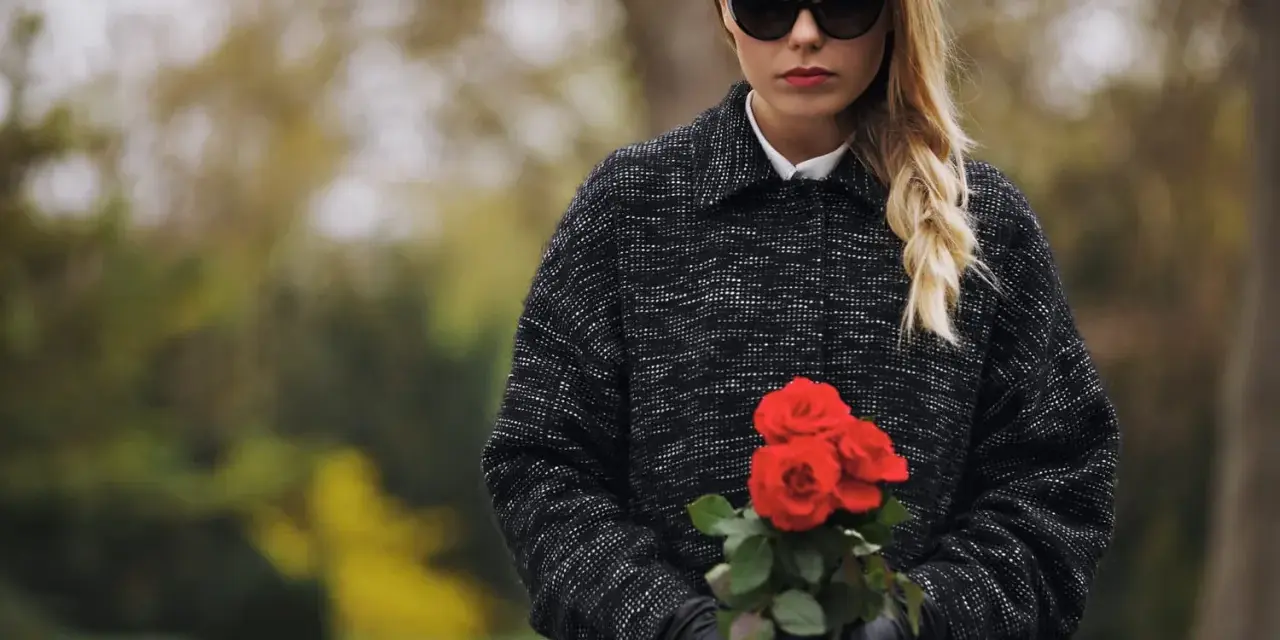 Une jeune femme, portant une tenue d'enterrement hiver, tient un bouquet de roses rouges. Ses lunettes de soleil cachent ses yeux, mais son expression est empreinte de tristesse.