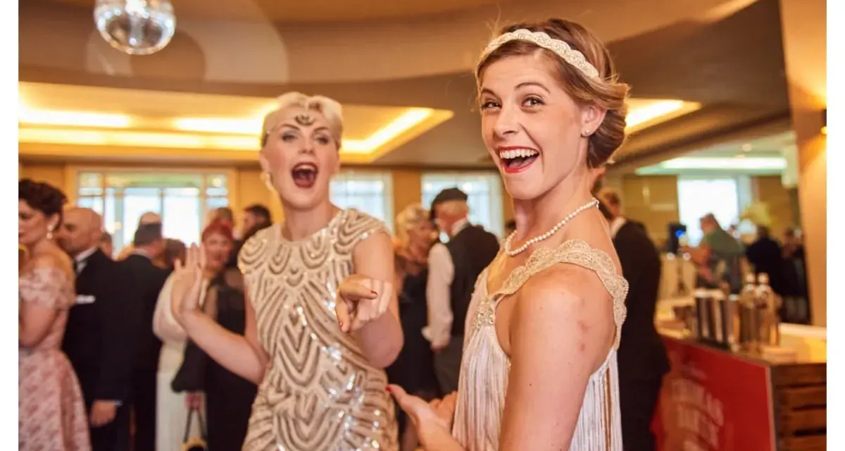 Deux femmes dansent, l'une porte une robe à paillettes dorées et un bandeau, l'autre une robe à franges, parfaites pour une tenue guinguette femme.