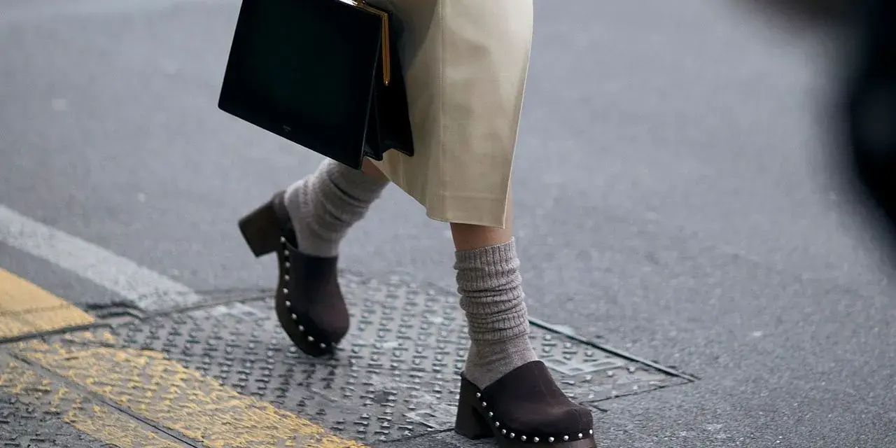 Une femme marche dans la rue, portant un sac noir et un élégant sabot femme marron clouté avec des chaussettes grises.