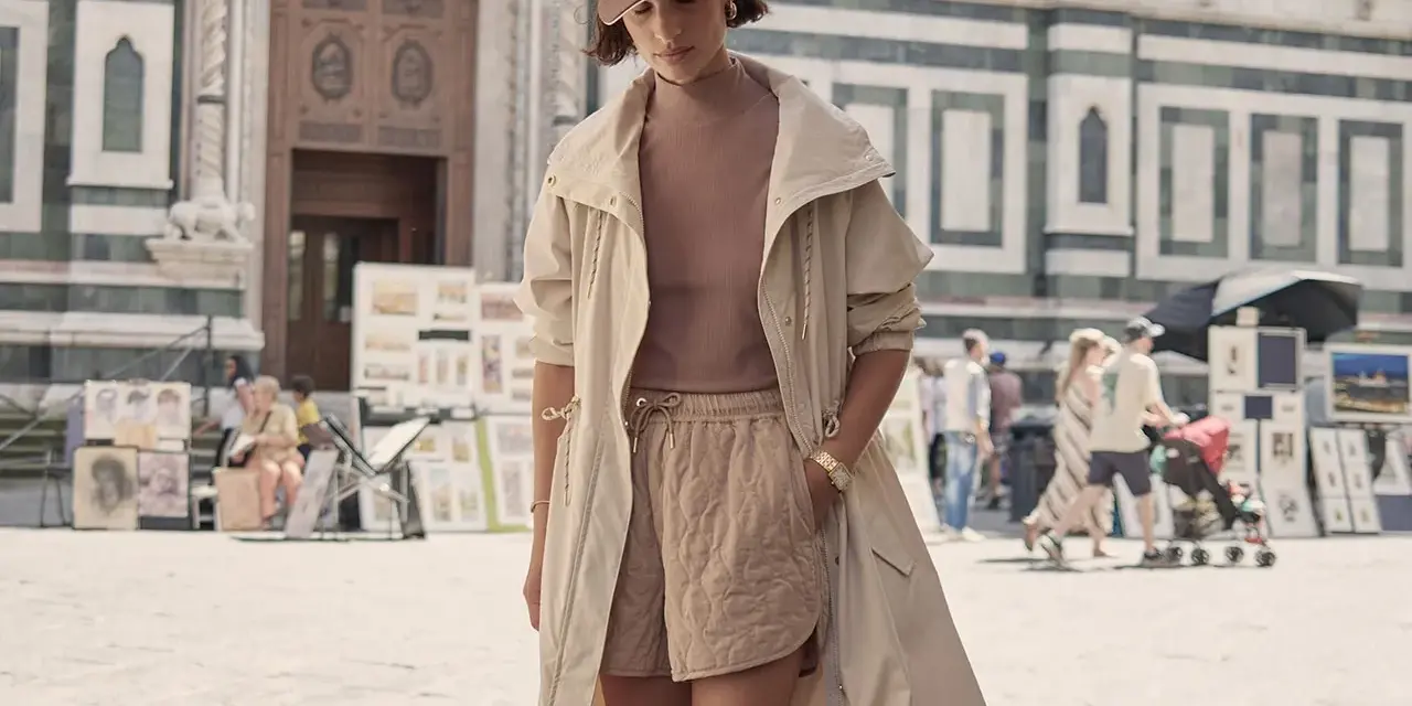 Une femme porte un ensemble athleisure chic : short matelassé, haut col roulé, veste longue et casquette. Elle se promène devant une cathédrale.