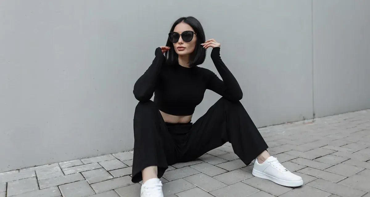 Une femme en tenue noire, crop top et pantalon large, assise sur des pavés. Idée de comment porter un pantalon large avec des baskets blanches.