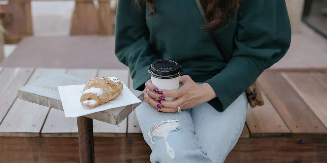Une femme en pull vert, jean déchiré, savoure un café et un croissant. Parfait pour un look décontracté.