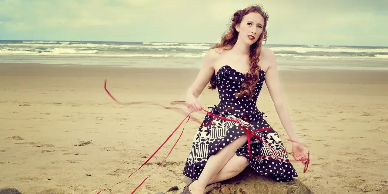 Jeune femme au style pin-up année 50, assise sur un rocher sur la plage, robe à pois, ruban rouge flottant au vent.