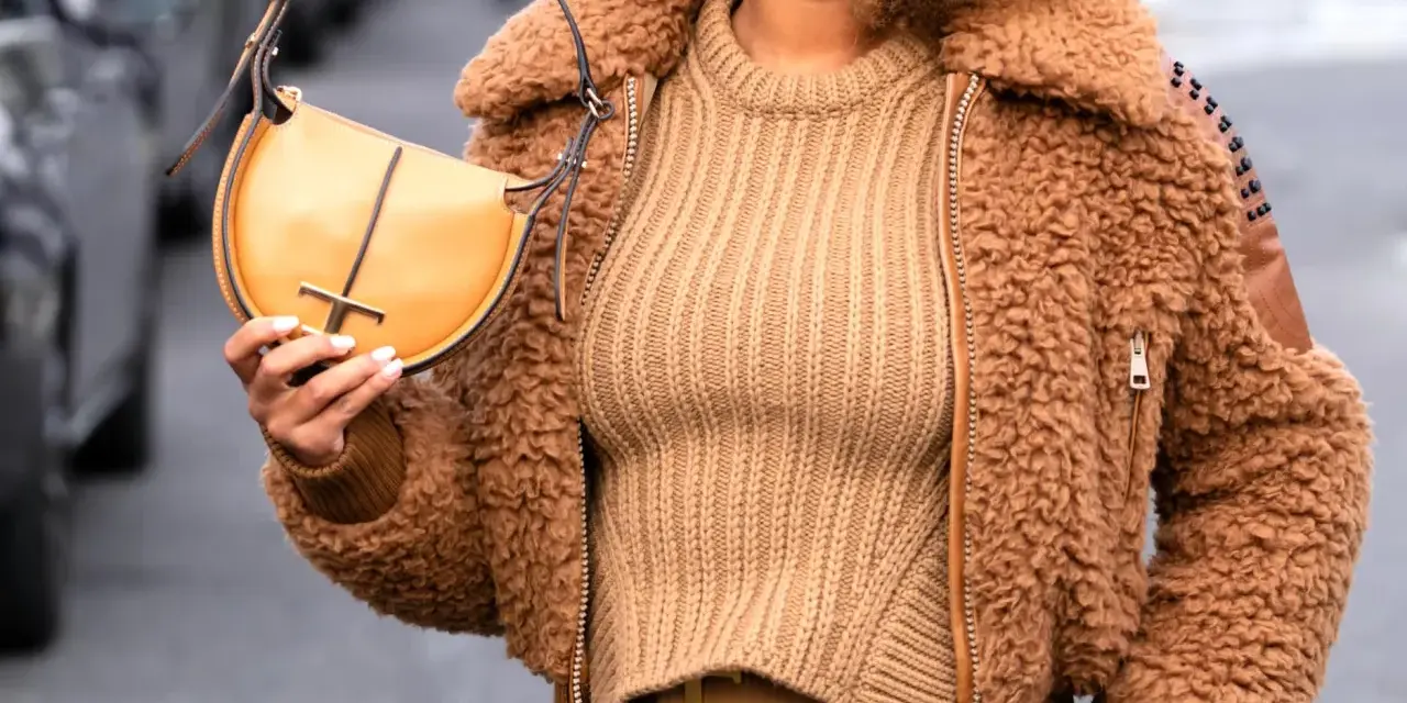 Une femme porte un pull beige côtelé, une veste en fausse fourrure marron et un sac à main jaune moutarde. Le jaune moutarde est une couleur qui va bien avec le beige.
