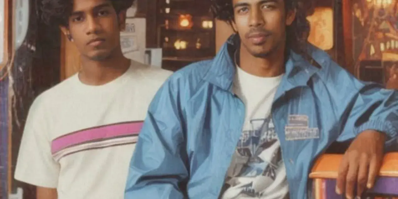 Deux jeunes hommes posent, arborant un style vestimentaire année 80 : t-shirt à rayures roses et veste bleue.