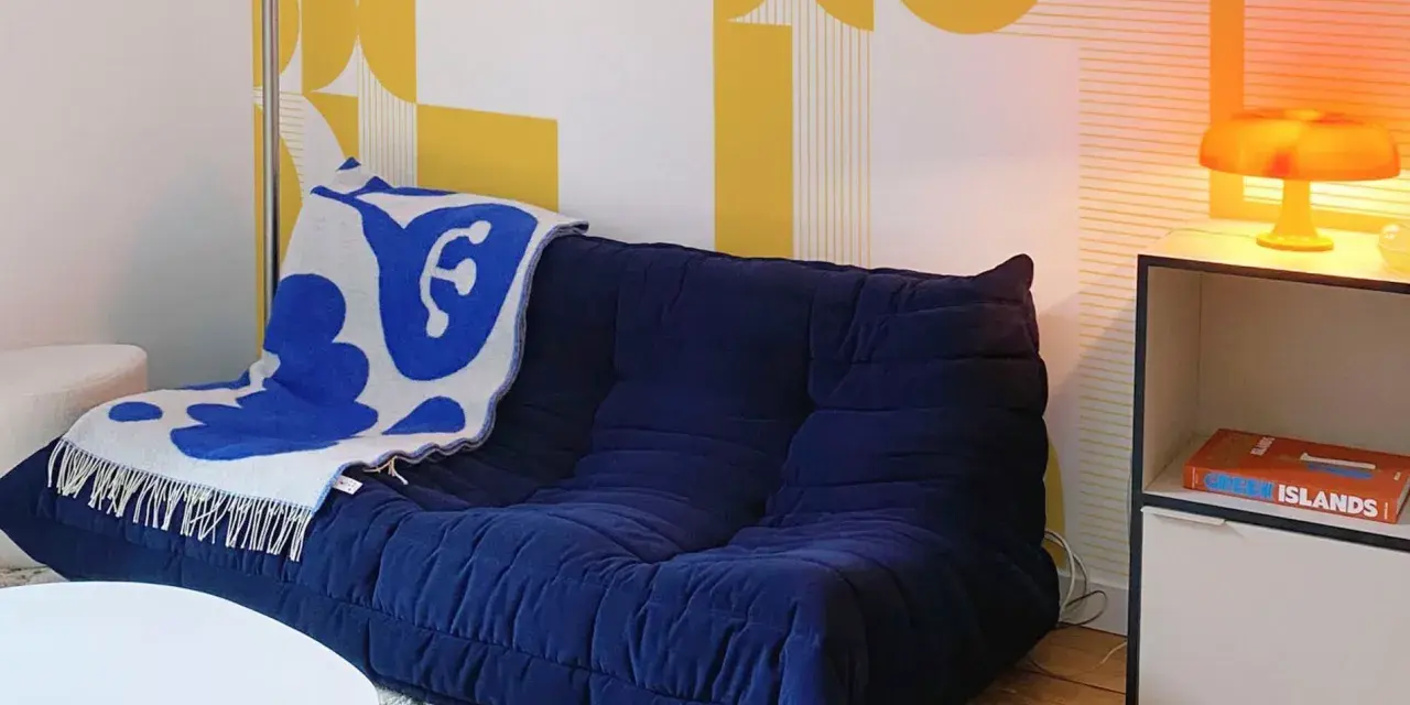 Canapé bleu profond avec plaid blanc et bleu, sur fond de papier peint jaune géométrique en **color blocking**. Lampe orange sur une étagère.