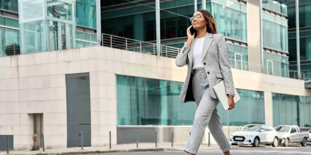 Femme en tenue d'entretien d'embauche d'été, tailleur gris, traversant la rue avec un ordinateur portable, parlant au téléphone.
