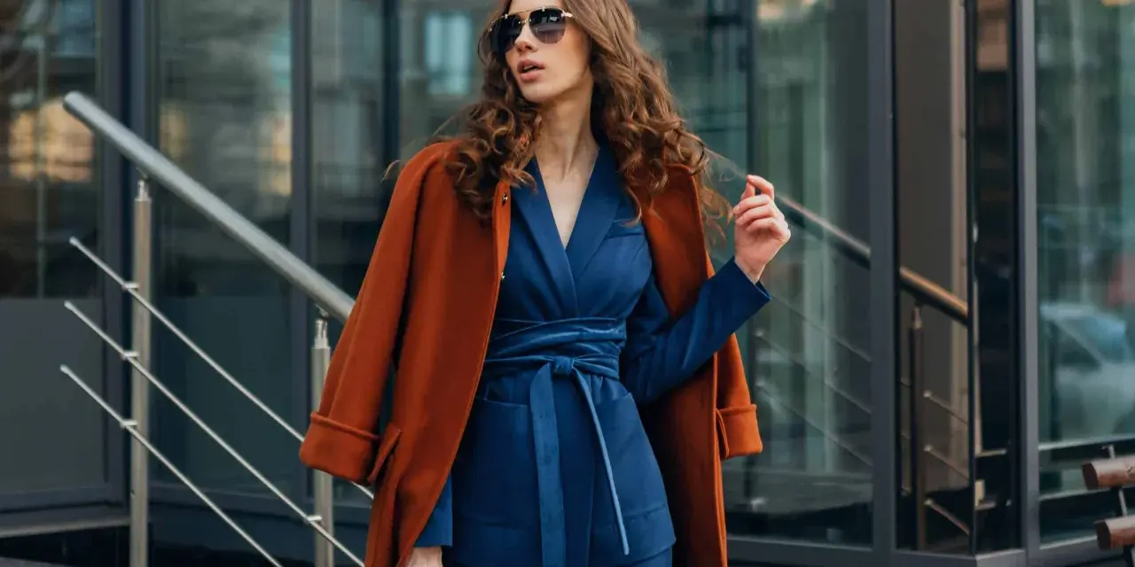 Une femme porte un gilet avec une robe bleu marine, un manteau orange sur les épaules, et des lunettes de soleil.