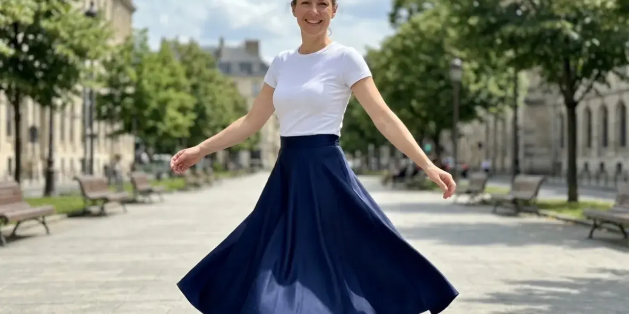 Une femme en talon et jupe évasée bleue danse joyeusement dans une rue parisienne bordée d'arbres.