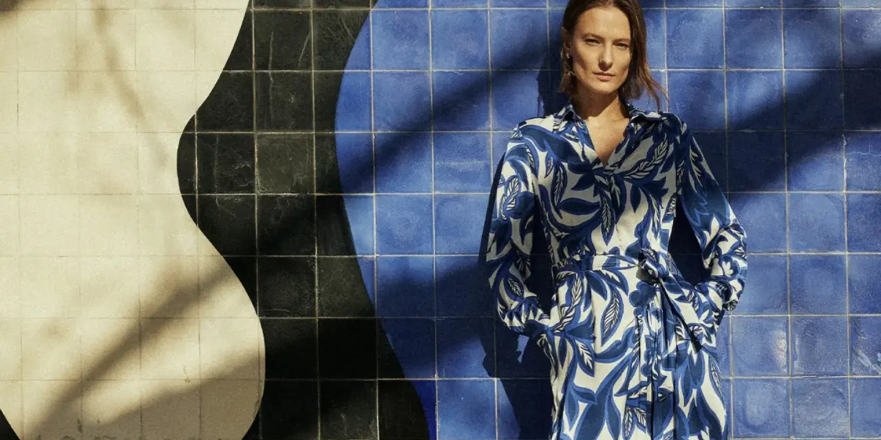 Une femme pose devant un mur de carreaux bleus et blancs. Sa robe à motifs bleus et blancs s'harmonise parfaitement avec le décor. Quelle couleur va bien avec le bleu vêtement ? Le blanc, évidemment !