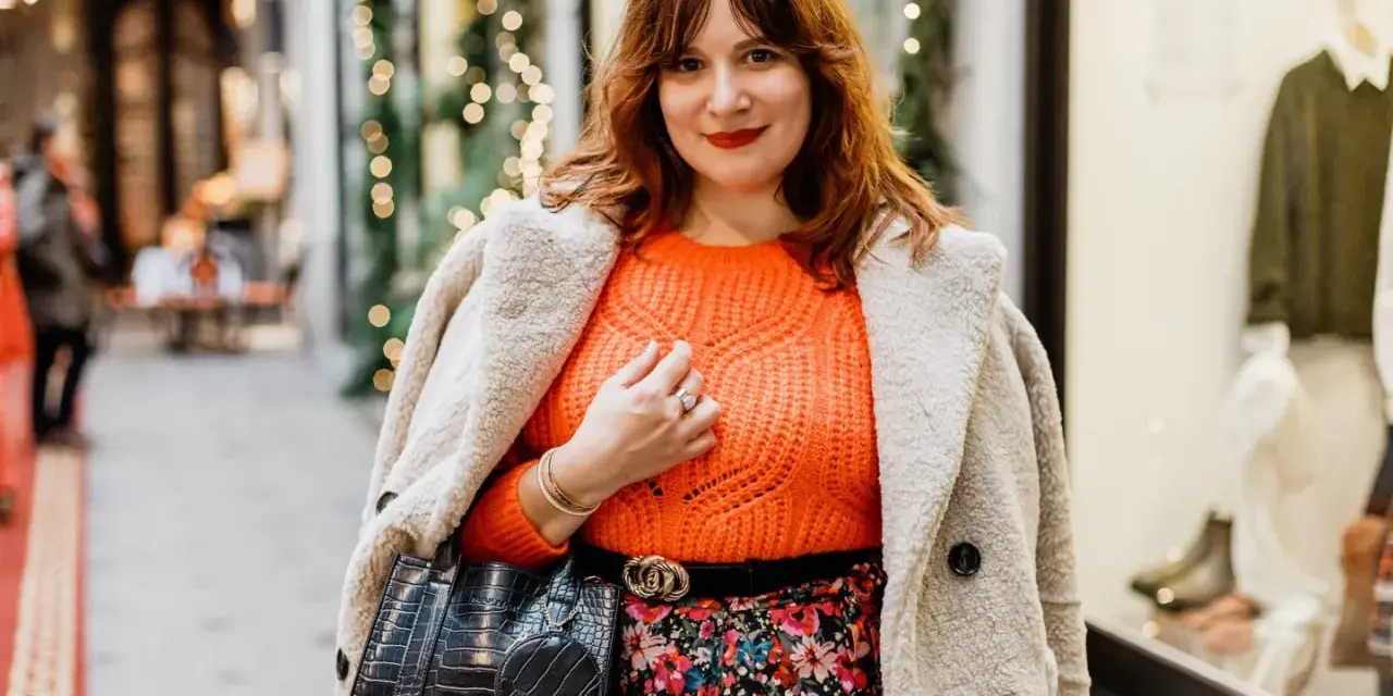 Une femme arbore une tenue avec pull orange vif, jupe fleurie et manteau douillet, prête pour une sortie festive.