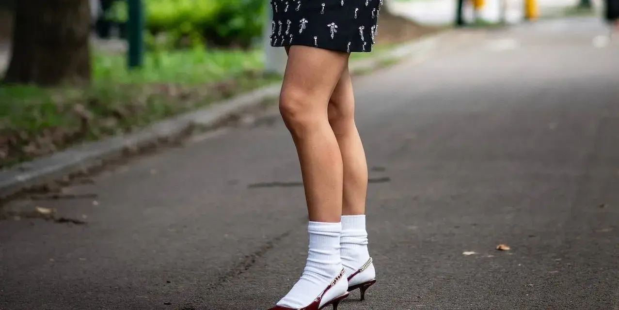Jambes portant une jupe ornée et des escarpins bordeaux avec une chaussette par dessus chaussure blanche.