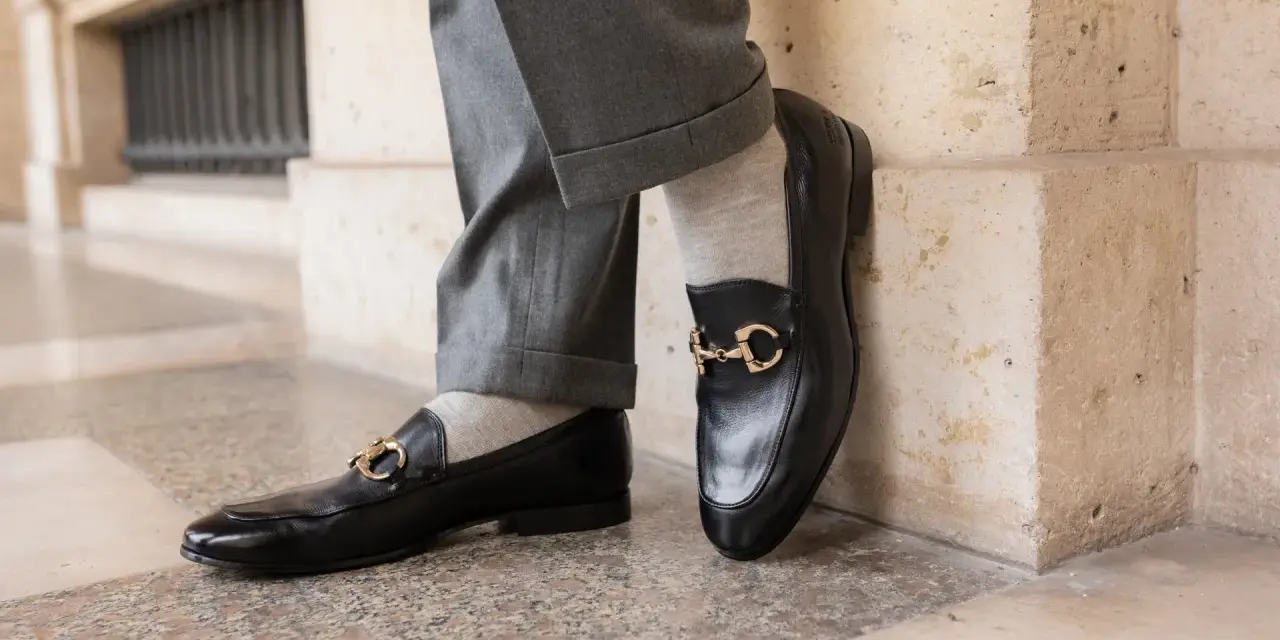 Un homme porte un pantalon gris retroussé et une chaussette mocassin noire avec une boucle dorée.