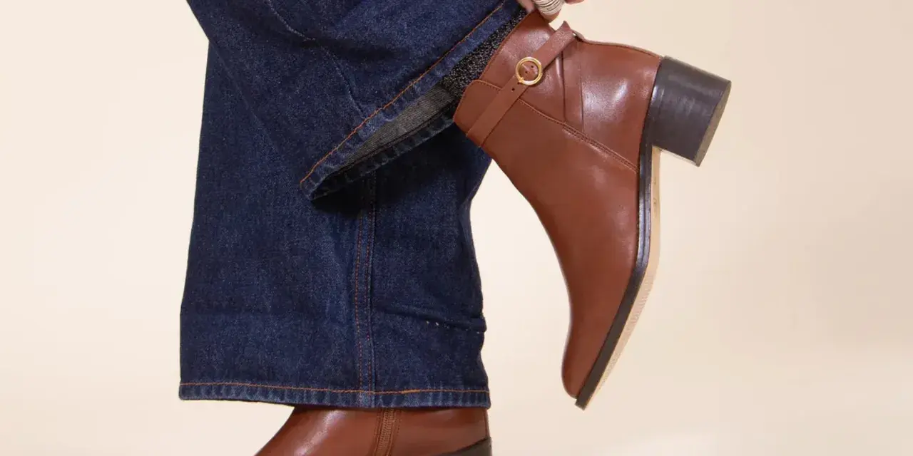 Une femme ajuste sa tenue avec bottines marron. Un jean évasé et un pull blanc complètent le look.