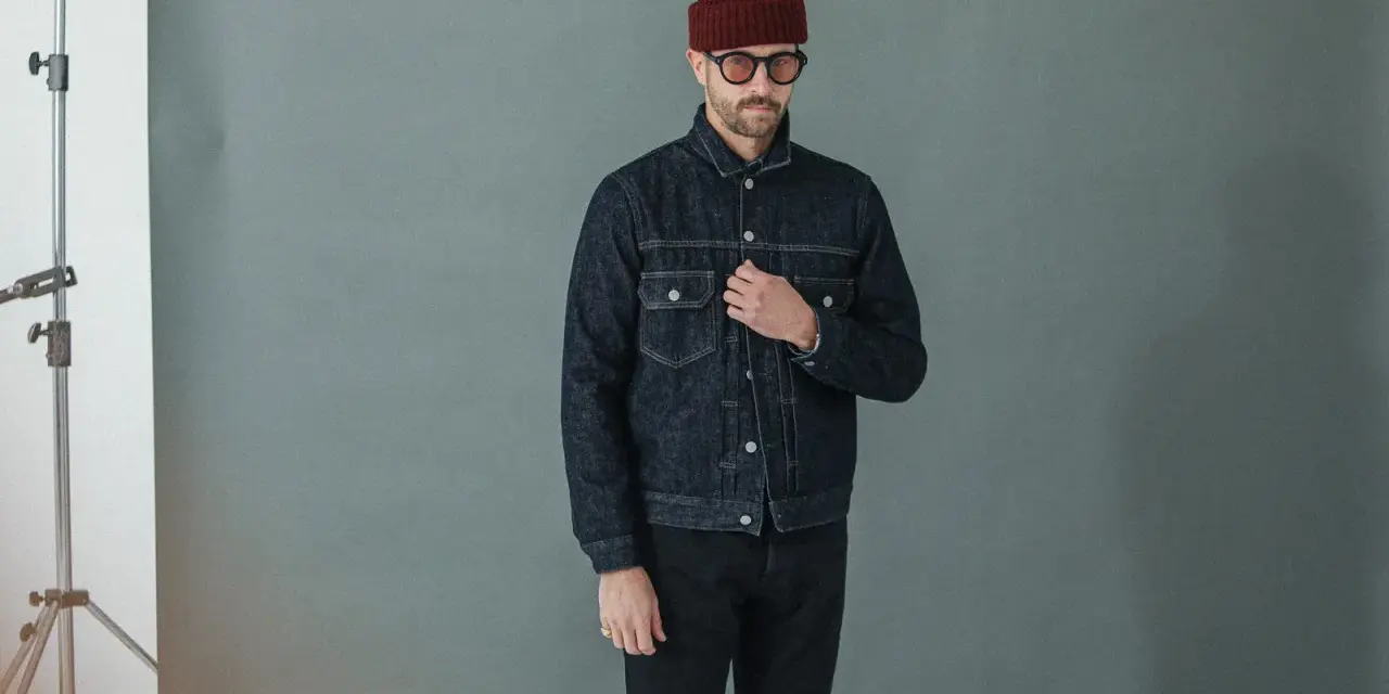 Homme portant une veste en jean sombre et un pantalon noir, bonnet bordeaux et lunettes rondes.