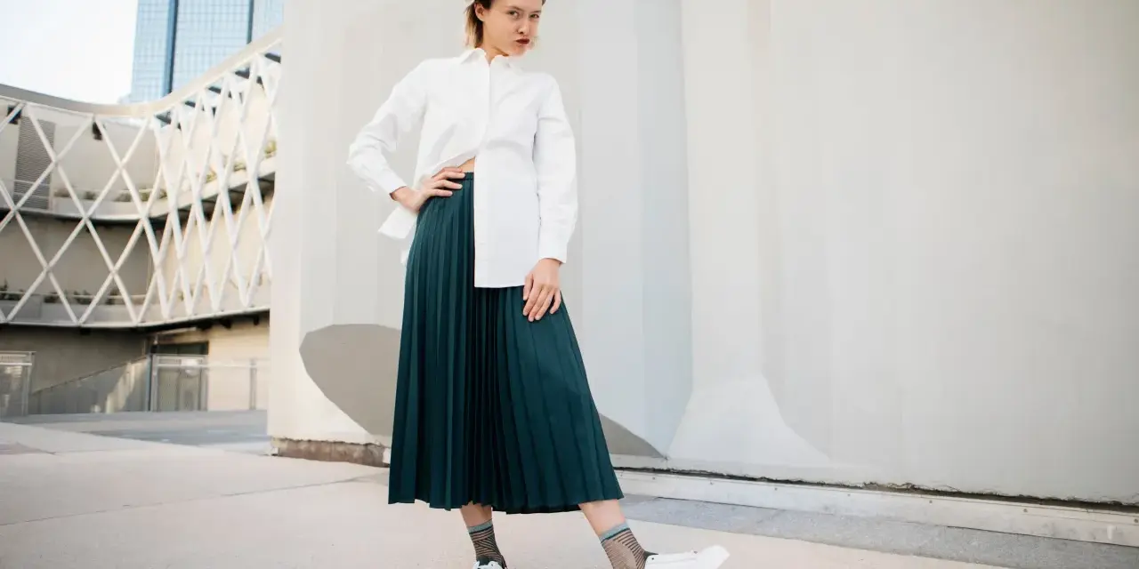 Une femme pose avec confiance, portant une chemise blanche et une élégante jupe plissée vert foncé.