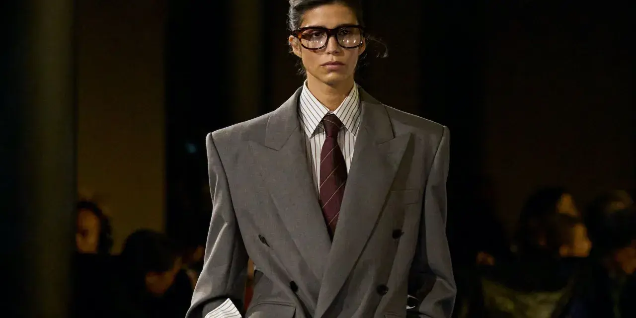 Mannequin au style collegienne, portant un costume gris oversize, chemise à rayures et cravate bordeaux. Lunettes imposantes.