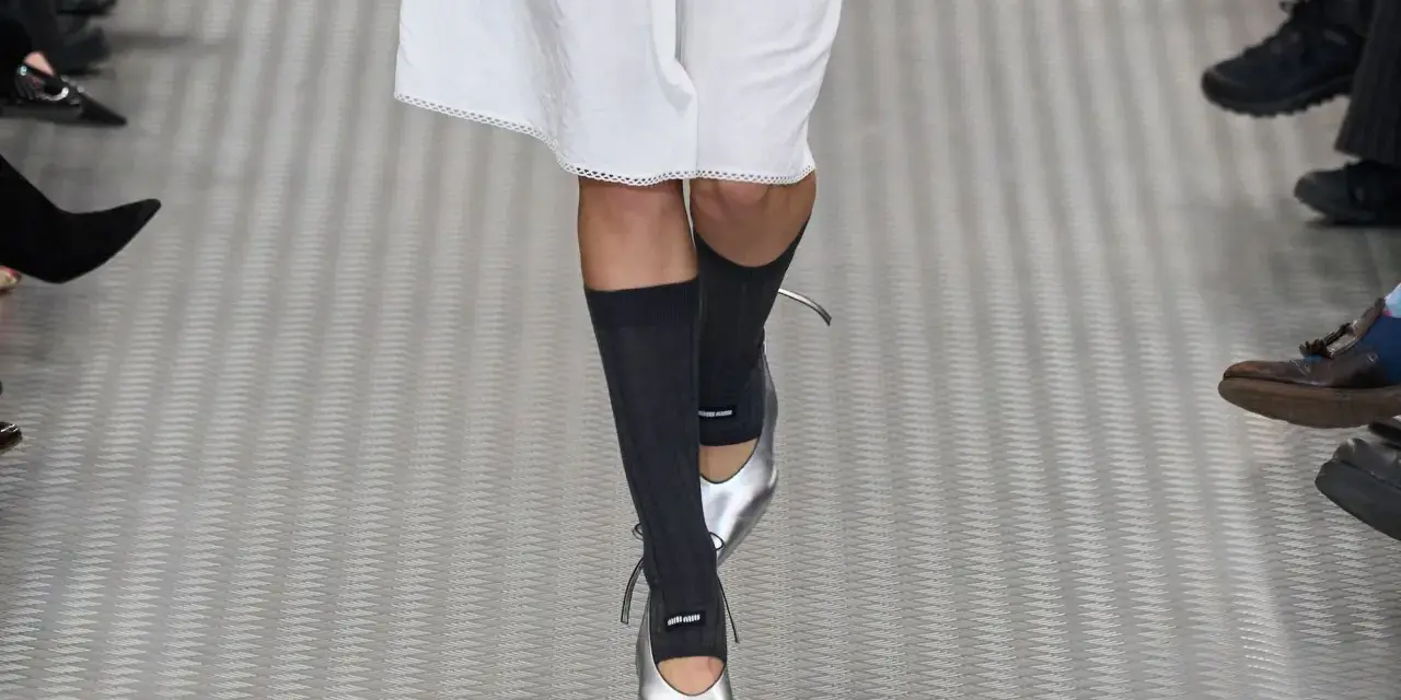 Une mannequin défile avec une tenue blanche et des chaussettes hautes grises, rehaussée par des chaussures argentées pointues.
