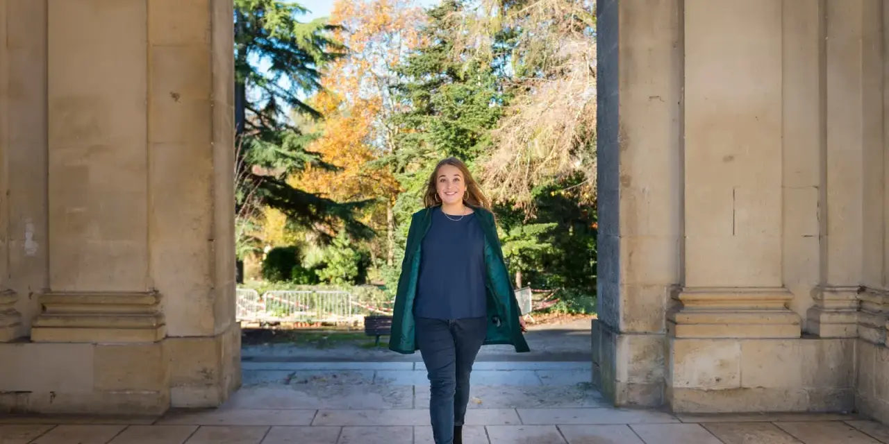 Une femme souriante marche sous une arche, portant un manteau vert et un haut bleu marine. L'automne colore les arbres en arrière-plan.