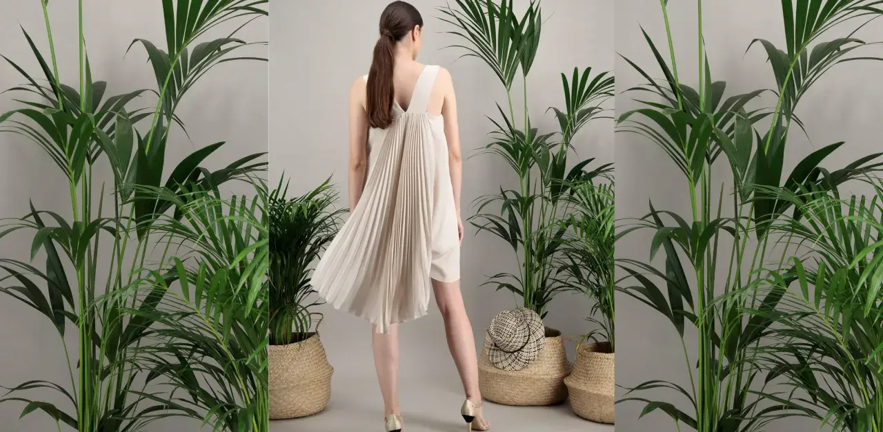 Une femme porte une robe plissée beige, parfaite pour une tenue mariage septembre. Elle est entourée de plantes vertes luxuriantes dans des paniers en osier.