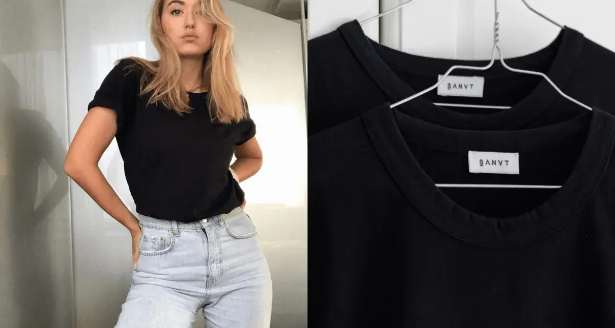 Une jeune femme porte une tenue avec t-shirt noir et jean clair. Deux t-shirts noirs identiques sont accrochés sur des cintres.